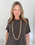 A woman with blonde hair wearing a dark brown short-sleeve t-shirt and a long gold vintage style link necklace, standing against a white wall.