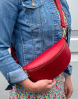 A person wearing a denim jacket is holding a red leather crossbody bag with a detachable strap.