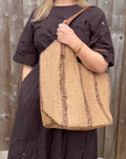 Woman wearing a brown dress holding a beige and brown raffia tote bag.