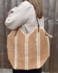 Woman wearing a white shirt, holding a large cream and beige raffia tote bag.