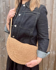 Woman wearing a denim jacket, holding a beige raffia crossbody handbag.