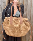 Woman wearing a black dress and denim jacket, holding a beige raffia shoulder handbag.