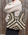 Woman wearing a white shirt holding a black and cream raffia tote bag.