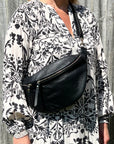 A person wearing a black large crossbody bum bag with an adjustable, detachable leather strap and multiple zip pockets.