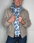 Woman wearing a beige jacket and blue jeans with a patterned scarf against a plain background