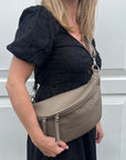 A person wearing a dark taupe large crossbody bum bag with an adjustable leather strap and multiple zip pockets.
