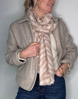 Woman wearing a beige jacket and a camel and beige scarf, and blue jeans against a plain background