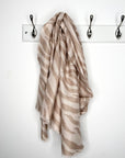 Beige and camel patterned scarf hanging on a white rack with three hooks against a white background.