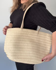 A person holding a cream-colored raffia tote bag with a PU handle strap.