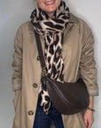 Person wearing a beige trench coat, blue jeans, and a brown crossbody bag with a leopard print scarf.