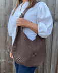 Woman wearing a white shirt and holding a brown woven crossbody handbag.