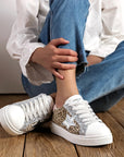 A person seated on wooden flooring wearing blue jeans and white leopard print & star lace-up trainers with a silver star and pale pink heel detail.