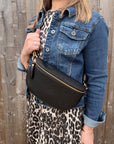 Woman wearing a denim jacket and leopard print skirt holding a large black crossbody/ waist bag against a wooden fence.
