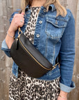 Woman wearing a denim jacket and leopard print skirt holding a large black crossbody/ waist bag against a wooden fence.