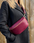 A person wearing a black coat is carrying a burgundy leather crossbody bag with gold hardware.
