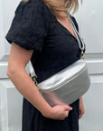 A woman wearing a black dress is holding a large silver leather crossbody bag with an adjustable strap.