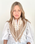 Woman wearing a white shirt and a light camel and cream patterned scarf against a plain background.