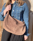 A woman wearing a denim jacket holding a mocha brown suede large crossbody swing bag with a detachable strap.