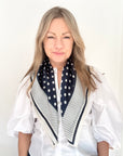 Woman wearing a white shirt and navy and cream patterned scarf against a plain background.