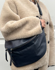 A woman wearing a beige coat is holding a navy blue leather crossbody bag with an adjustable strap and silver hardware.