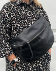 Woman wearing a black leather crossbody bag with silver hardware and an adjustable strap over a leopard print coat.