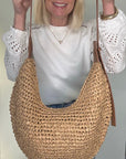 Woman wearing a white top, blue jeans, holding a raffia and tan woven crossbody handbag.