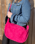 A person wearing a denim jacket is holding a raspberry large suede leather crossbody swing bag with a detachable strap.