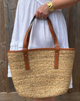 Person holding a straw tote bag with brown straps against a wooden background