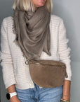 a woman wearing a white jumper and a grey scarf, holding a dark taupe suede crossbody/ waist bag.