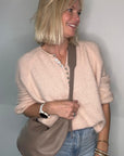 Woman wearing a beige sweater and blue jeans, holding a taupe crossbody/ shoulder bag