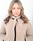 Woman wearing a beige coat with a taupe scarf against a white background