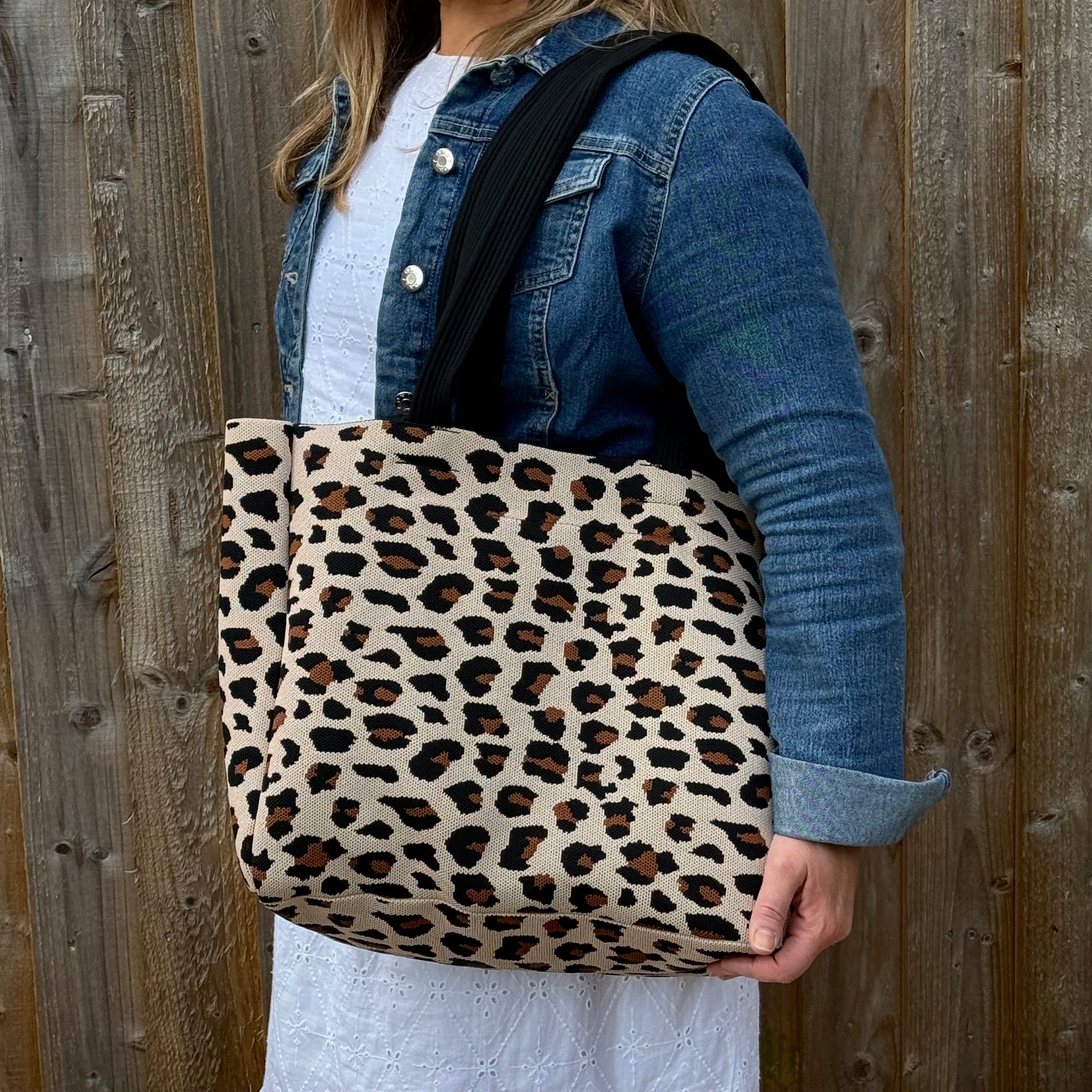 Person holding a leopard print tote bag against a wooden fence.