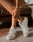 A pair of grey and gold leopard print heel trainers with white laces, worn by a person seated on a sofa.