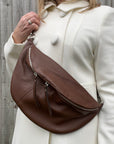 A woman wearing a brown large crossbody bum bag with an adjustable, detachable leather strap and multiple zip pockets.