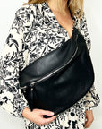 A person wearing a black XL crossbody/bum bag with silver hardware, in front of a white background with floral print clothing.