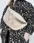 A person wearing a cream-colored large crossbody bum bag with an adjustable, detachable leather strap and multiple zip pockets, carried over the shoulder.