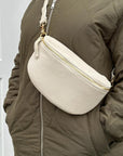 a woman wearing a large cream crossbody/ waist bag with and adjustable strap and gold hardware.