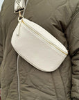 a woman wearing a large cream crossbody/ waist bag with and adjustable strap and gold hardware.