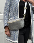 A woman wearing a silver crossbody waist bag with an adjustable strap, standing in front of a white background.