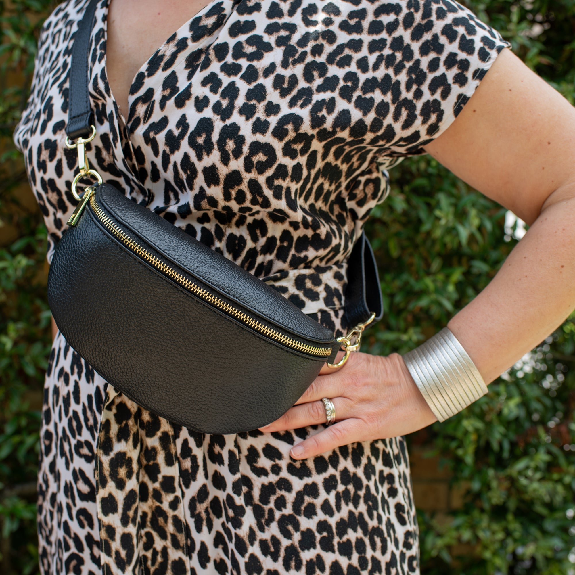 A person wearing a leopard print dress carrying a black leather waist bag with gold hardware.