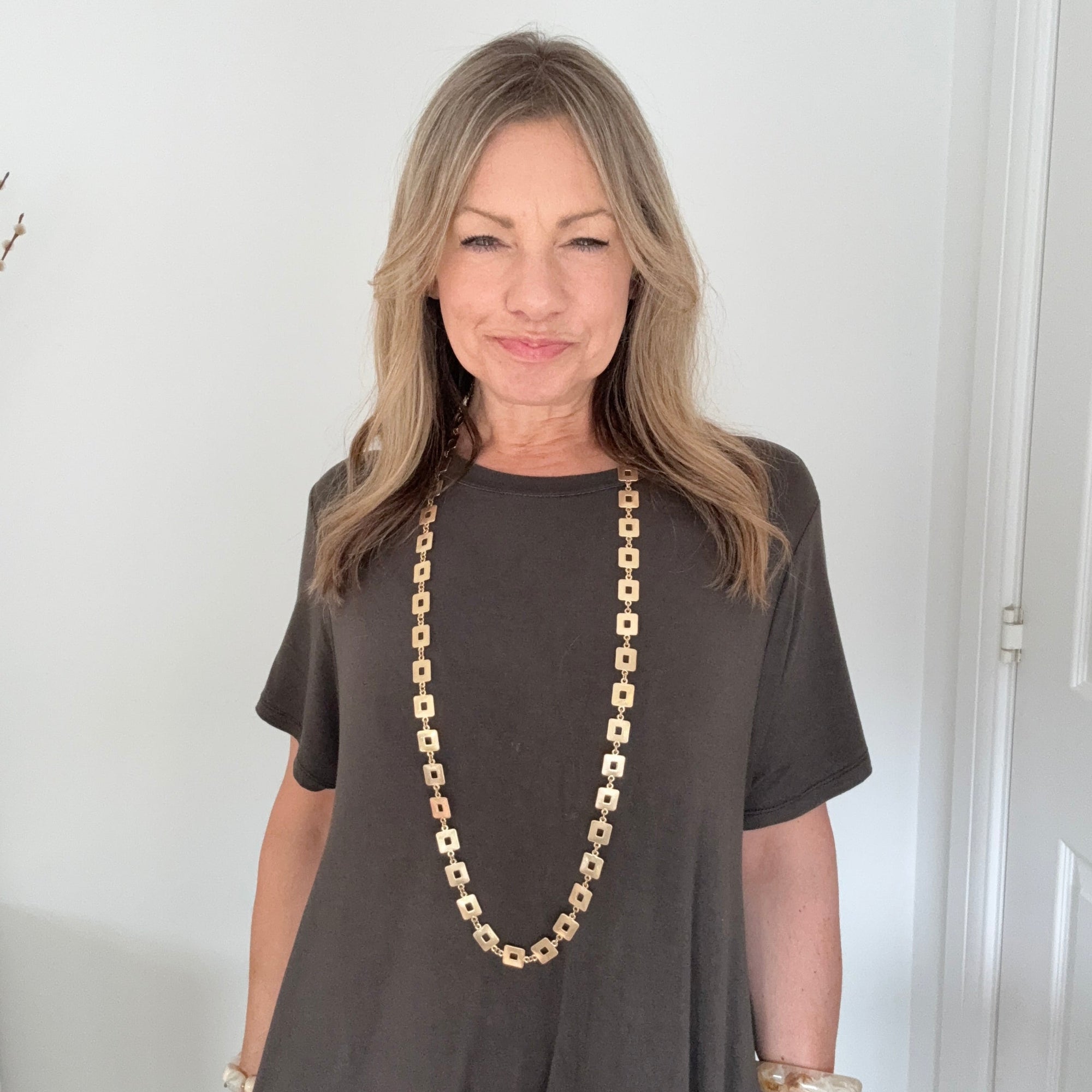 A woman with blonde hair wearing a dark brown short-sleeve t-shirt and a long gold vintage style link necklace, standing against a white wall.