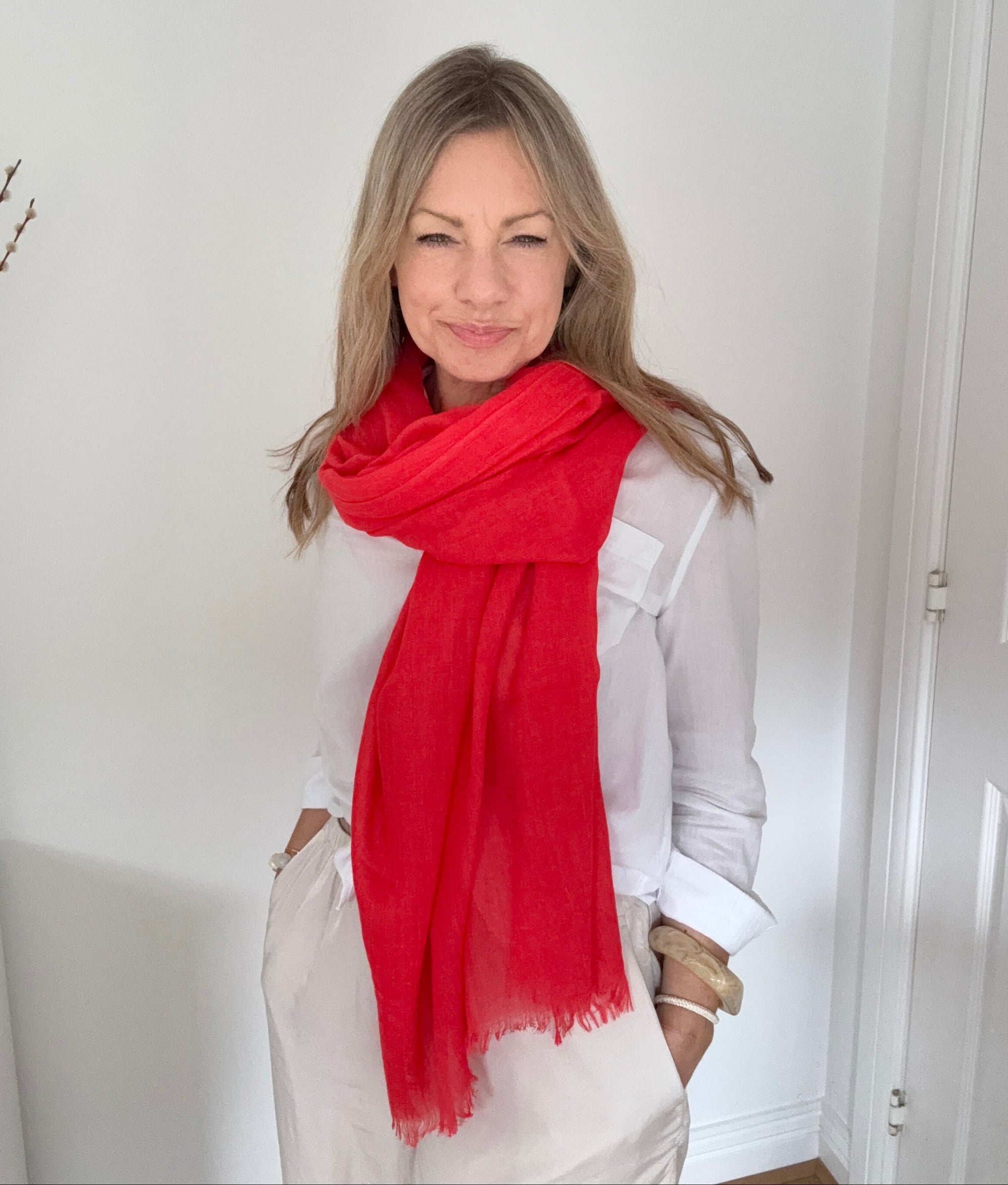 A woman with blonde hair wearing a white long-sleeve shirt and white pants, wrapped in a bright red scarf, standing in a white room.