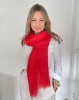 A woman with blonde hair wearing a white long-sleeve shirt and white pants, wrapped in a bright red scarf, standing in a white room.