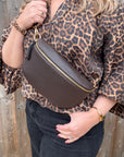 Person wearing a leopard print top and holding a large dark brown leather waist crossbody bag 