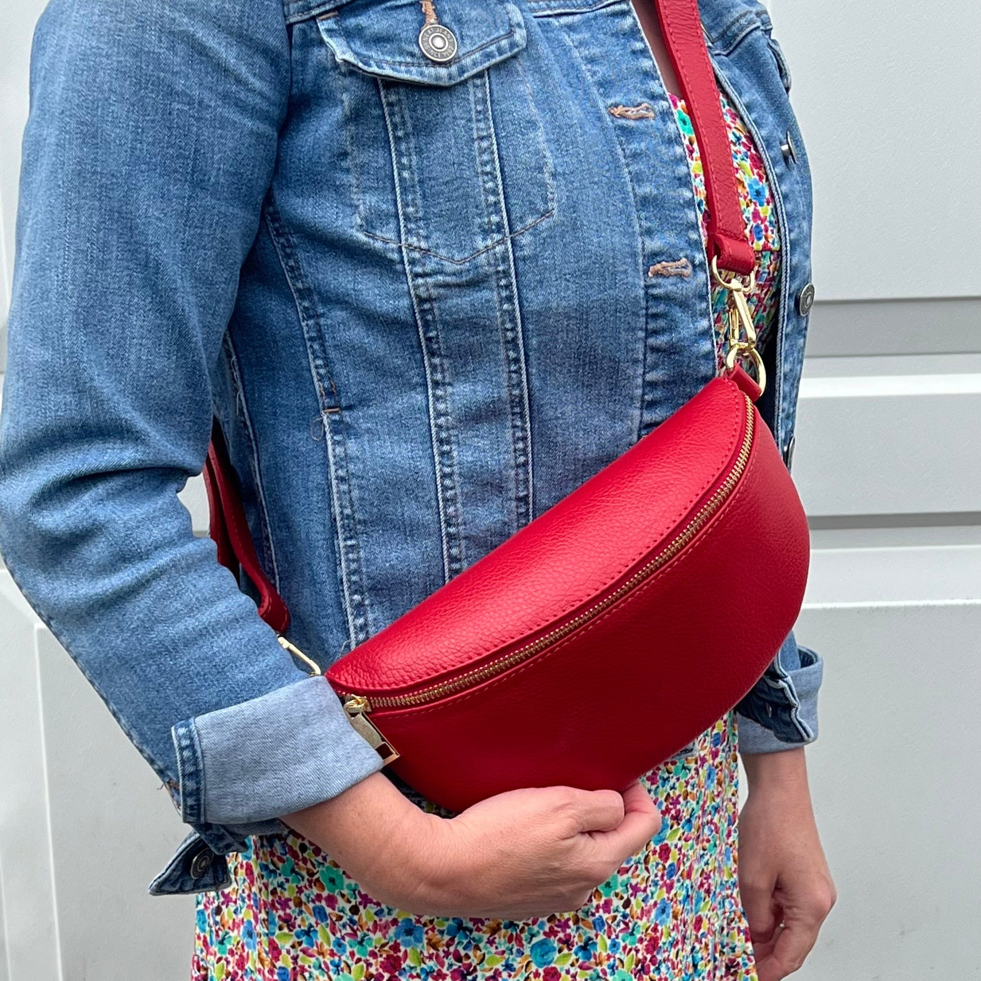 A person wearing a denim jacket is holding a red leather crossbody bag with a detachable strap.