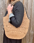 Woman wearing a denim jacket and black skirt, holding a beige and black woven raffia shoulder handbag.