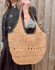 Woman wearing a denim jacket and black skirt, holding a beige and black woven raffia shoulder handbag.