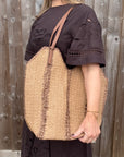 Woman wearing a brown dress holding a beige and brown raffia tote bag.