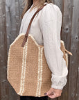 Woman wearing a white shirt, holding a large cream and beige raffia tote bag. 