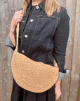 Woman wearing a denim jacket, holding a beige raffia crossbody handbag. 